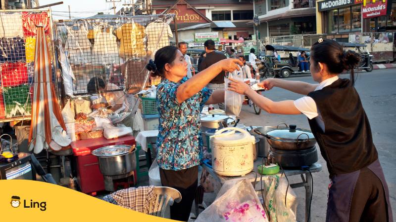 Mädchen steht bei einer Köchin am Street Food Stand in Thailand und möchte Essen auf Thai bestellen