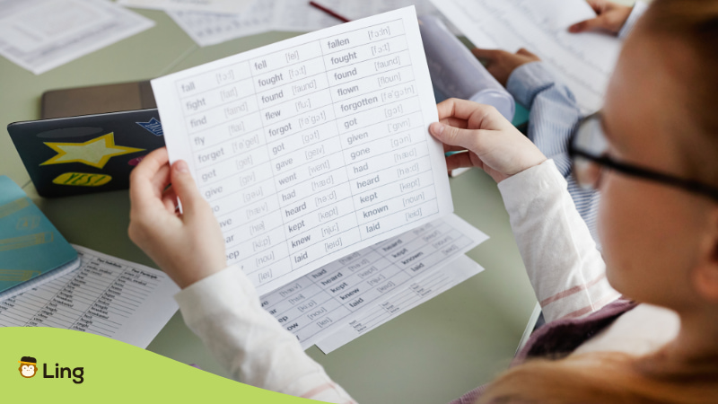 A Student Holding A Printed Verb Conjugation Chart Featuring Verbs In Different Tenses And Pronunciations, With Study Materials Spread Across The Table