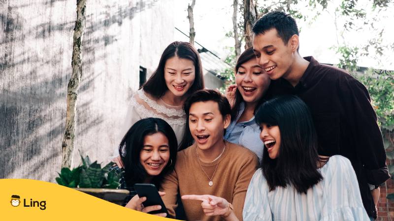 Gruppe von 6 thailändischen Freunden treffen sich und schauen gemeinsam lachend in ein Smartphone