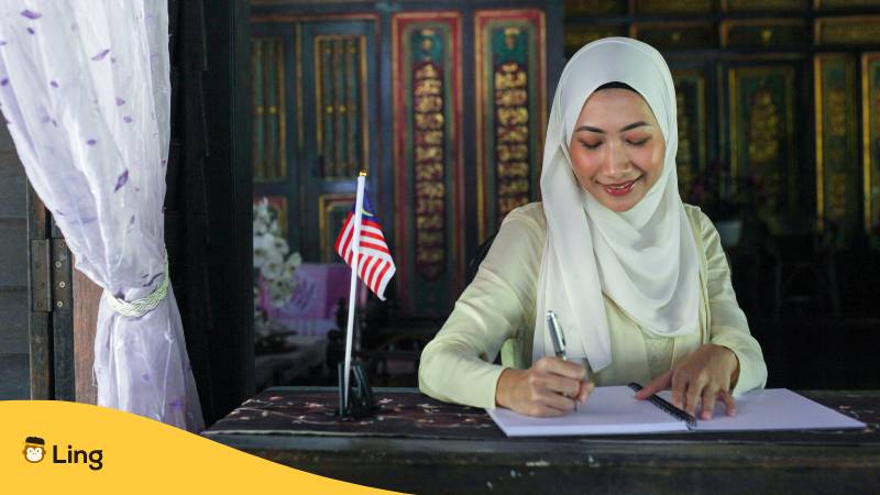 Frau sitzt an einem Schreibtisch mit malaysischer Flagge und schreibt mit dem Jumi und Rawi Alphabet
