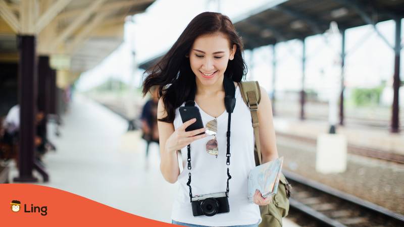 Reisende Frau mit Kamera um den Hals und einer Karte und Handy lernt unterwegs Thai mit der Ling-App