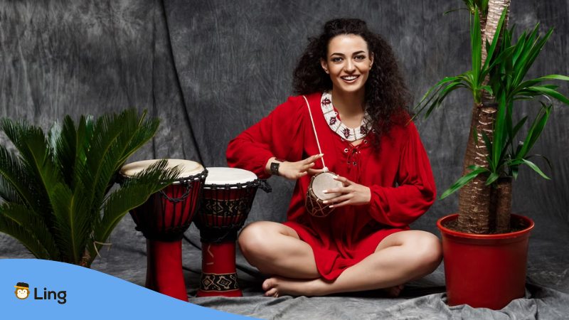 A photo of a beautiful girl playing a drum in tropical plants over a grey wall.