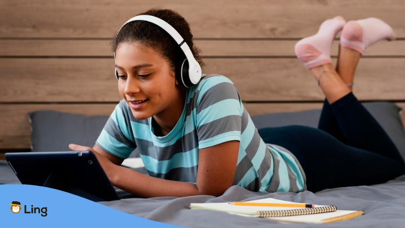 A girl Thai language learner with a tablet and headphones on the bed.