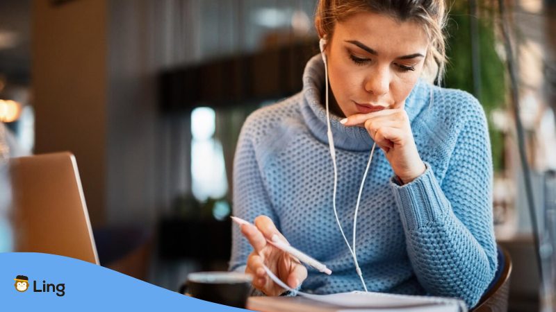 A young casual businesswoman working in a cafe and going through paperwork while listening to podcasts in Thai over earphones.