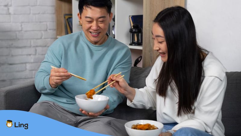 A photo of a Korean couple sitting on a couch eating Korean fried chicken with their chopsticks.