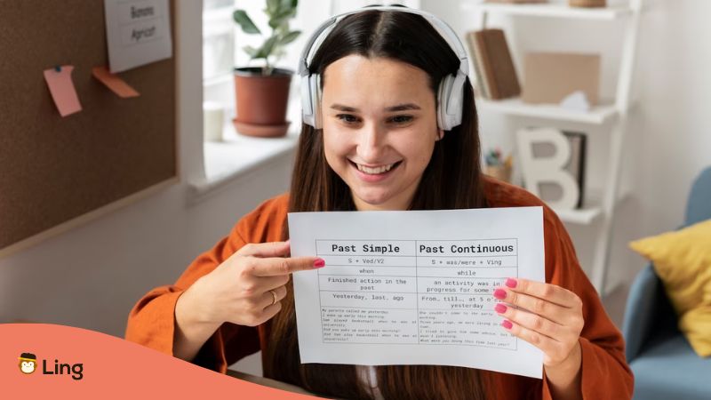 A smiling femal language learner with headphones showing verb tenses on a paper.