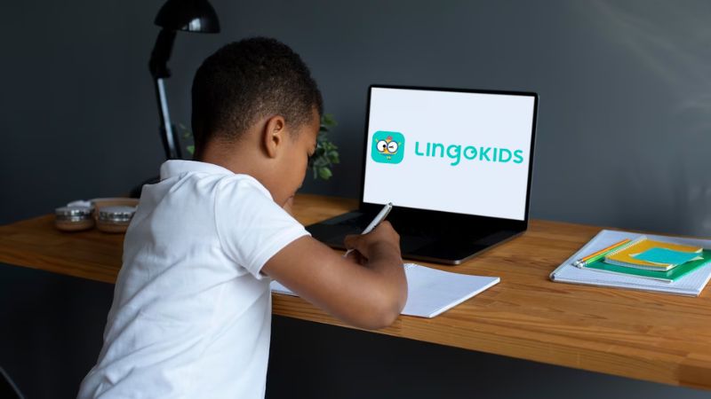 A photo of a boy writing on his notebook on a table and a laptop infront with Lingokids on the screen.