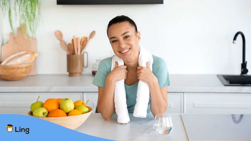 A photo of a smiling woman in her kitchen just finished working out with a towel around her neck.