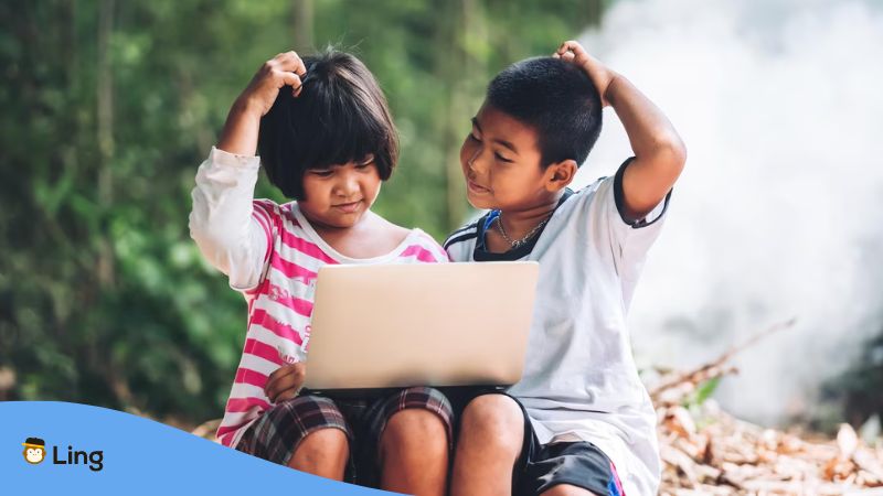 A photo of two kids sitting outdoors scratching their heads while using a laptop.