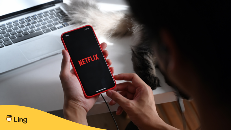 man browsing netflix on phone with a cat and laptop on his table 