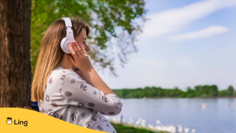 Blonde Frau sitzt am See und hat Kopfhröer auf und hört dänische Lieder und nutzt die Ling-App um die dänische Sprache lernen