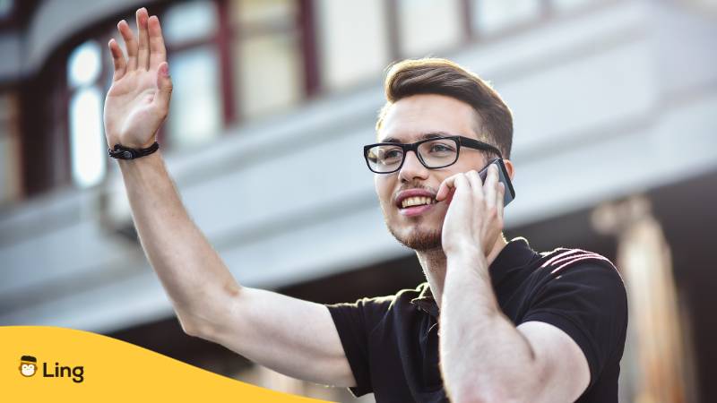 Blonder Mann mit Brille telefoniert mit seinem Smartphone und winkt jemanden zu um ihn auf Ukrainisch zu begrüßen
