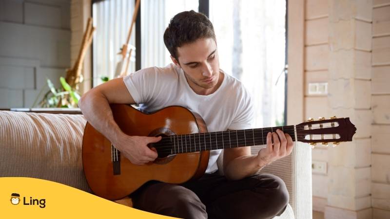 Junger Mann sitzt mit seiner Gitarre auf einem Sofa und übt litausche Lieder mit der Ling-App