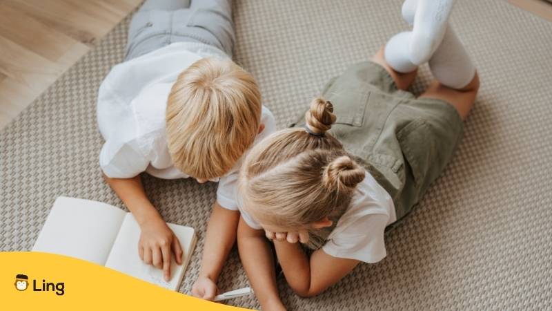 Ein litauischer Junge und Mädchen liegen auf dem Teppich und schauen sich faszinierende Namen in litauischer Sprache für Jungen und Mädchen in einem Buch an. Entdecke litauische Namen für deinen Nachwuchs mit der Ling-App.