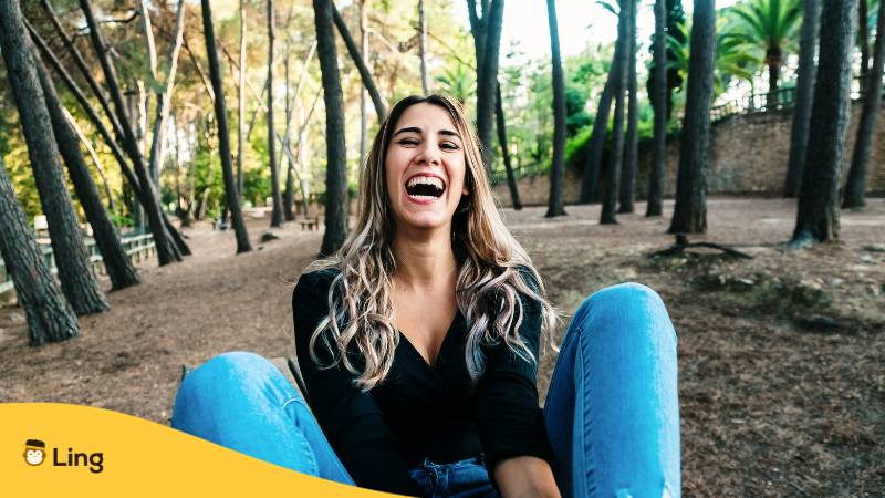 Junge Frau sitzt im Wald auf dem Boden und lacht über litauische Witze, die sie mit der Ling-App kennengelernt hat