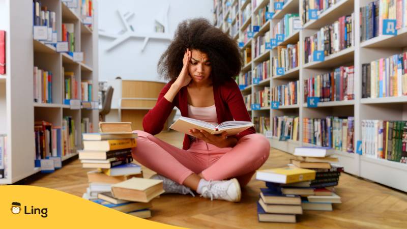 Junge Frau mit vollem lockigen Haaren sitzt zwischen Bücherstapeln und Regalen und fragt sich, ist Malaysisch schwer zu lernen?