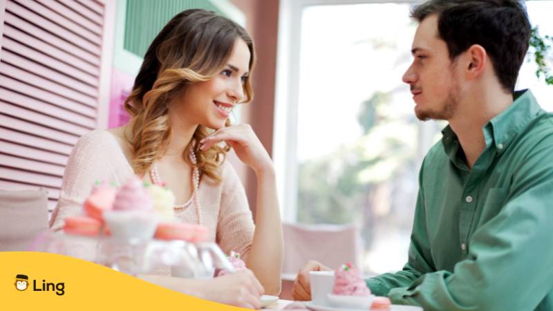 Date sitzt gemeinsam im Café. Sie legt ihr Kinn auf ihre Hand und lächelt ihn charmant an. Erfahre alle über Liebeswörter und Sätze auf Litauisch für Dates mit der Ling-App.