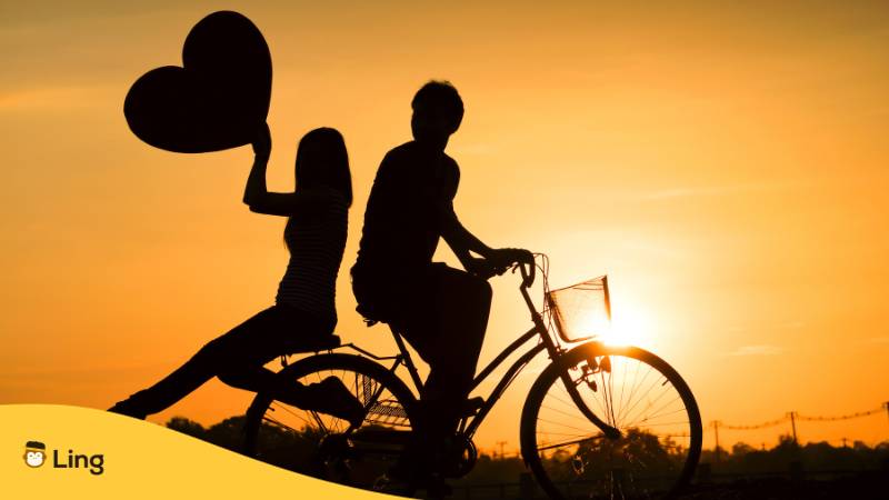 Schwarzer Umriss von Paar auf Fahrrad während Sonnenuntergang. Er fährt Fahrrad, sie sitzt mit Rücken zu ihm auf Gepäckträger und hält ein großes Herz in die Luft. Erfahre alles über Liebeswörter und Sätze auf Litauisch mit der Ling-App.