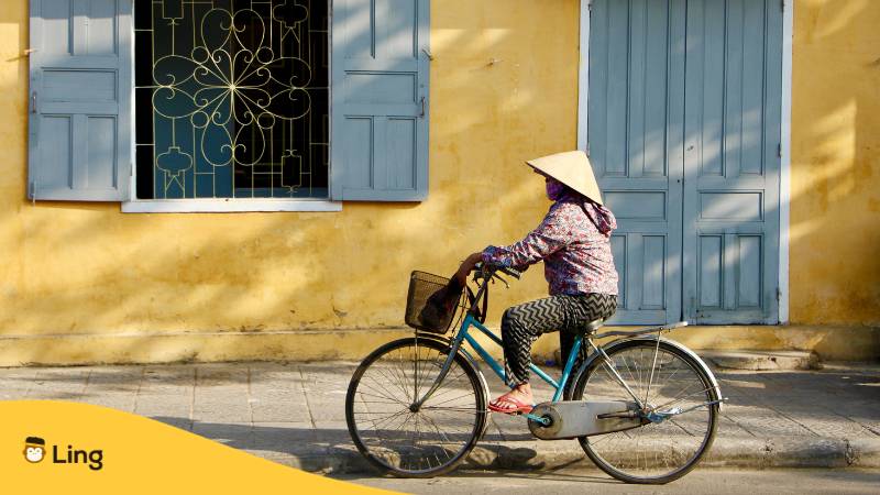 Vietnamesin fährt mit dem Fahrrad in Vietnam und trägt einen Hut im asiatischen Stil. Erfahre mit der Ling-App, ist Vietnamesisch schwer zu lernen.