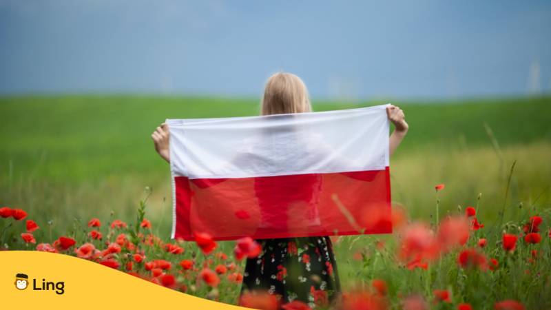 Blonde Polin hält polnische Flagge über Schultern und Rücken und blickt in die Ferne, während sie auf eine Mohnblumenwiese steht. Lerne wichtige polnische Sätze mit der Ling-App.