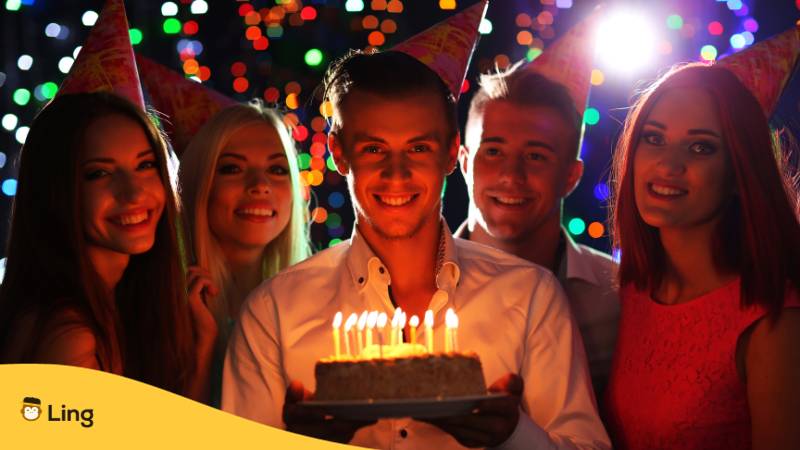 Junger Mann, der Geburtstag hat, hebt seine Geburtstagstorte mit Kerzen, wobei seine Freunde um ihn alles Gute zum Geburtstag auf Litauisch sagen.