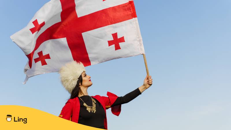 Georgisches Mädchen mit Nationalflagge von Georgien in den Händen auf blauem Himmelhintergrund. Georgische Kultur Lebensstil. Frau in Papakha und rotem Kleid. Lerne 30+ einfache georgische Sätze. Eine grundlegende Liste mit der Ling-App.
