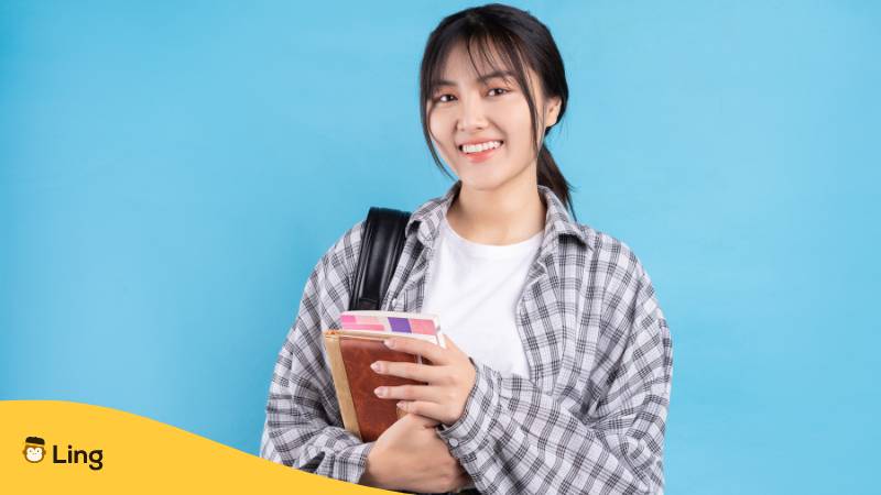 Asiatische Studentin mit freundlichem Gesichtsausdruck auf blauem Hintergrund. Lerne das thailändische Alphabet mit dem ultimativen Guide zur thailändischen Schrift mit der Ling-App.

