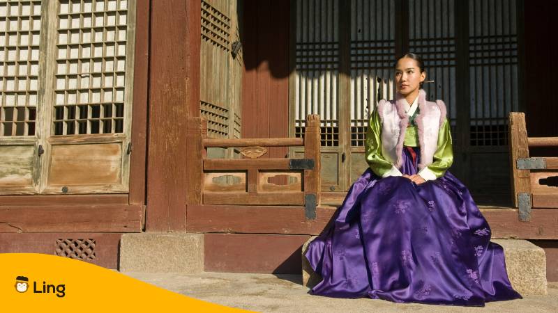 Junge Koreanerin trägt den koreanischen Nachnamen und sitzt in traditioneller Kleidung vor einem Haus. 
