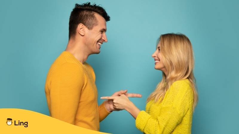 Mann und Frau mit gelben Oberteilen zeigen lächelnd mit dem Finger auf sich, nachdem sie sich die gleiche Frage gestellt haben. Lerne die 5 besten Fragewörter auf Kroatisch mit Ling!