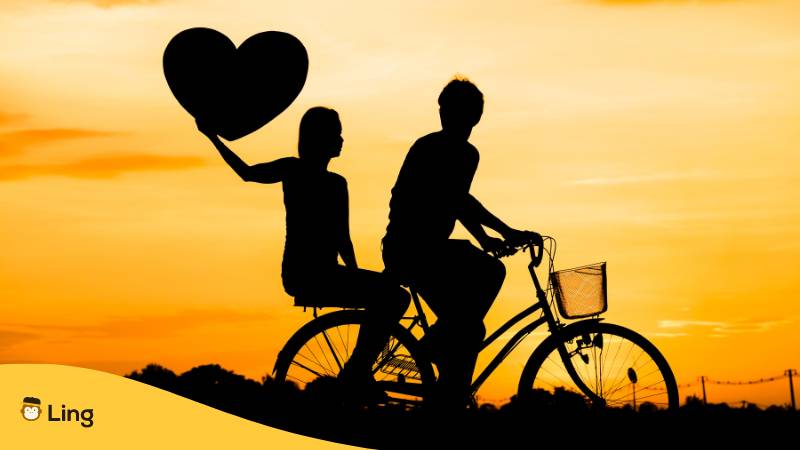 Silhouette eines Paars auf einem Fahrrad, mit einem großen Herz dabei. Lerne 25 Verzaubernde Liebesvokabeln auf Albanisch.

