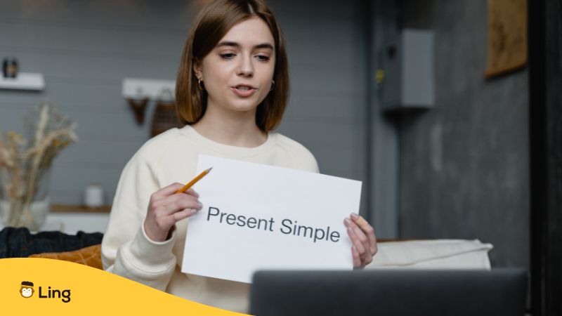 Woman holding a paper sign that says 'Present Simple' while teaching a grammar lesson