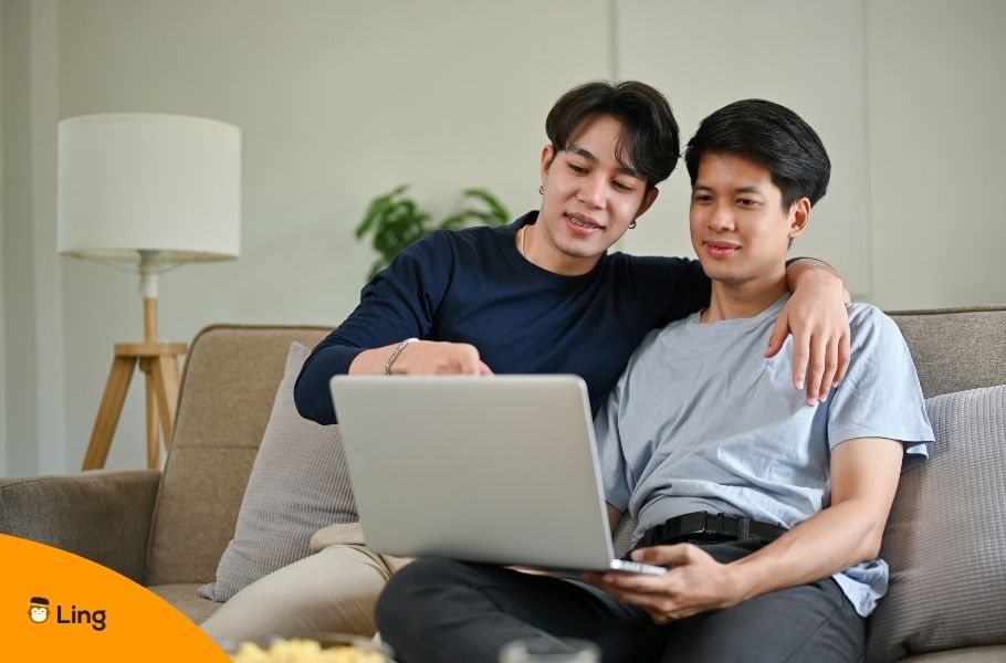 Smiling Asian Gay Couple Hugging And Using Laptop While Sitting Close Together On Sofa In Living Room At Home